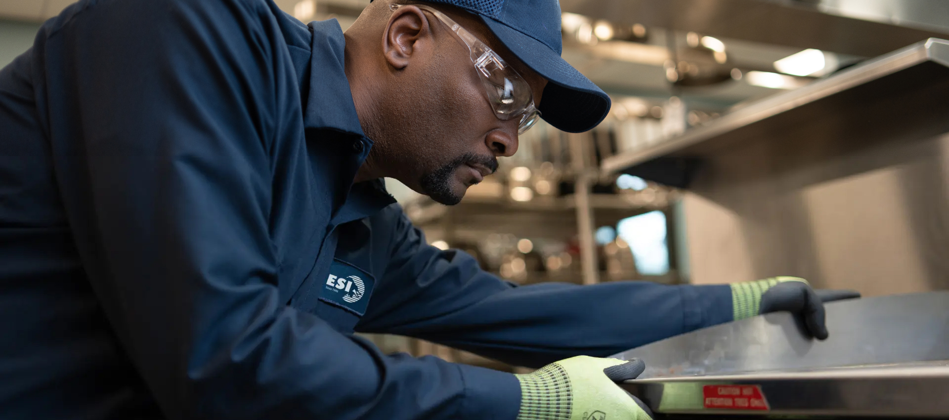 ESI technician in a branded navy work shirt and cap leaning over a stainless steel commercial range to service it
