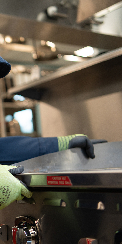 ESI technician in a branded navy work shirt and cap leaning over a stainless steel commercial range to service it