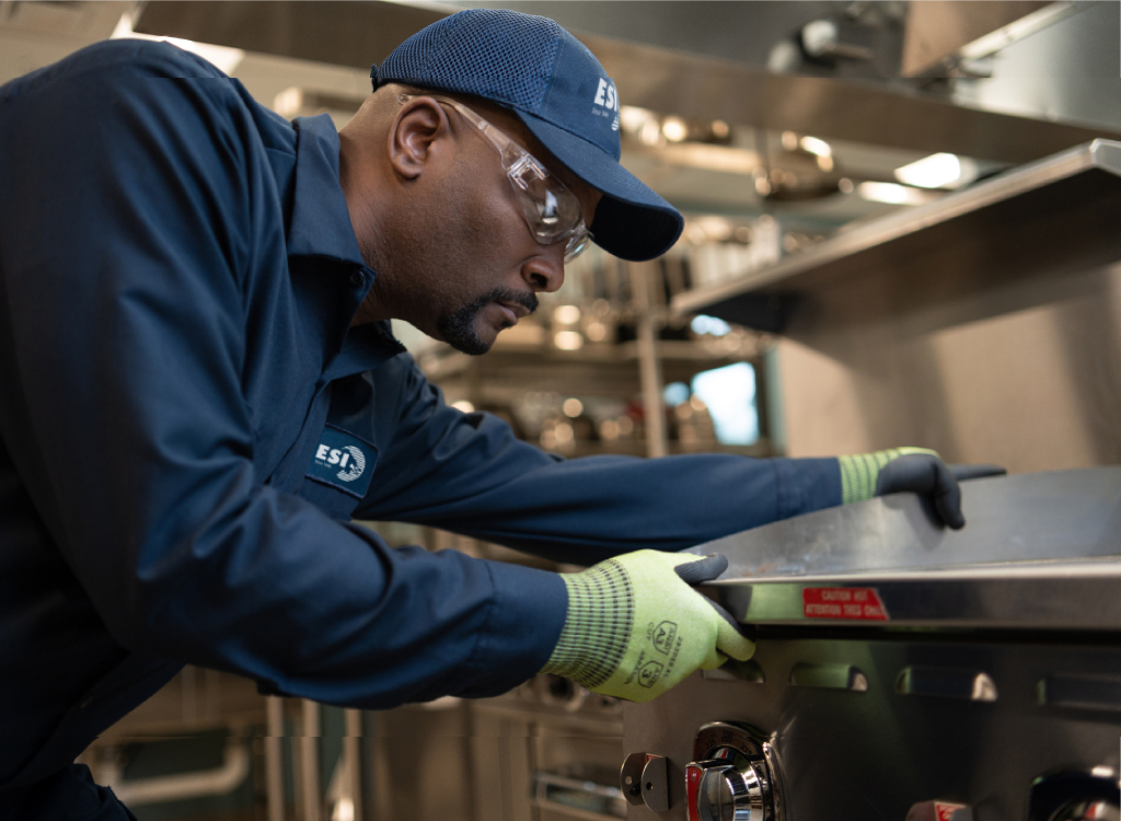 ESI technician in a branded navy work shirt and cap leaning over a stainless steel commercial range to service it