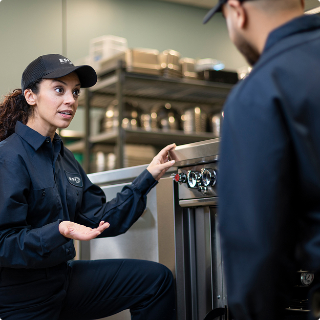 Two ESI technicians in branded caps and navy uniforms talking beside a commercial appliance during hands-on training