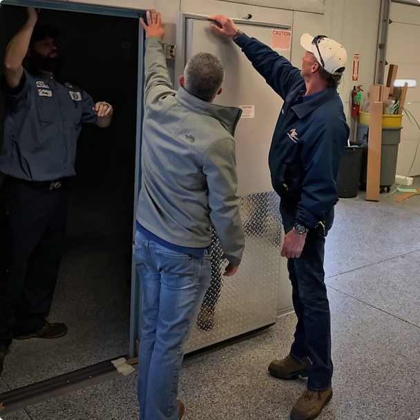Three ESI technicians examining the open door of a stainless steel walk-in cooler in the service shop