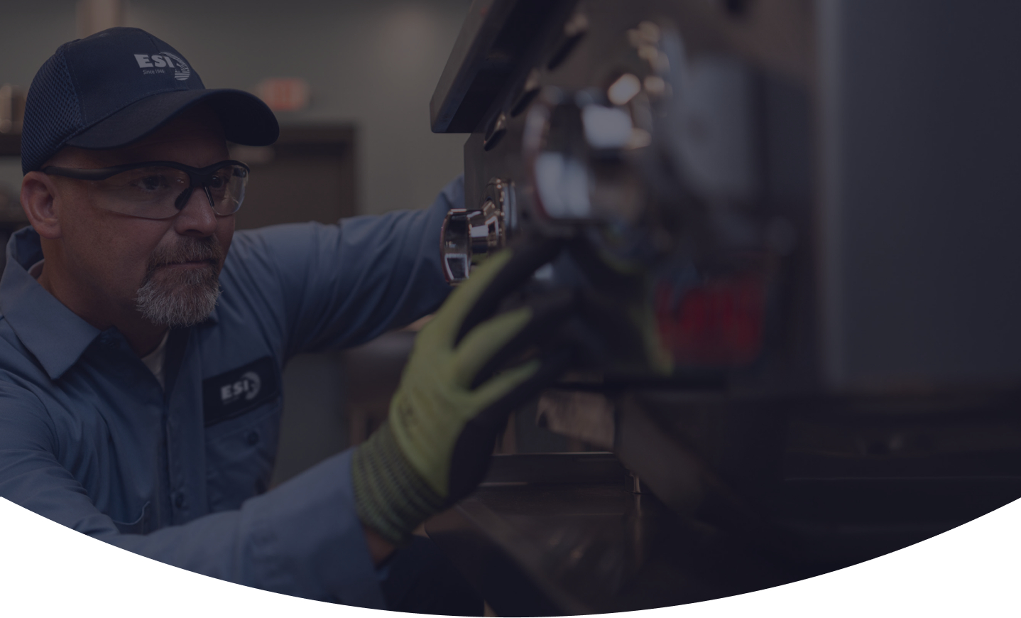 ESI technician in a branded cap and safety glasses reaching into the burner assembly of a piece of commercial cooking equipment