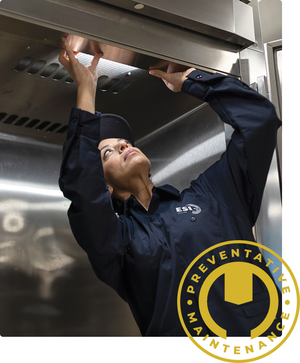 ESI technician reaching up under a stainless steel commercial oven, overlaid with a gold Preventative Maintenance badge