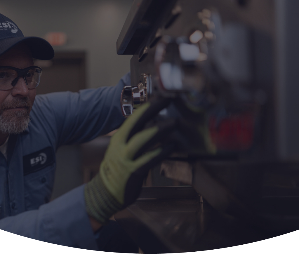 ESI technician in a branded cap and safety glasses reaching into the burner assembly of a piece of commercial cooking equipment