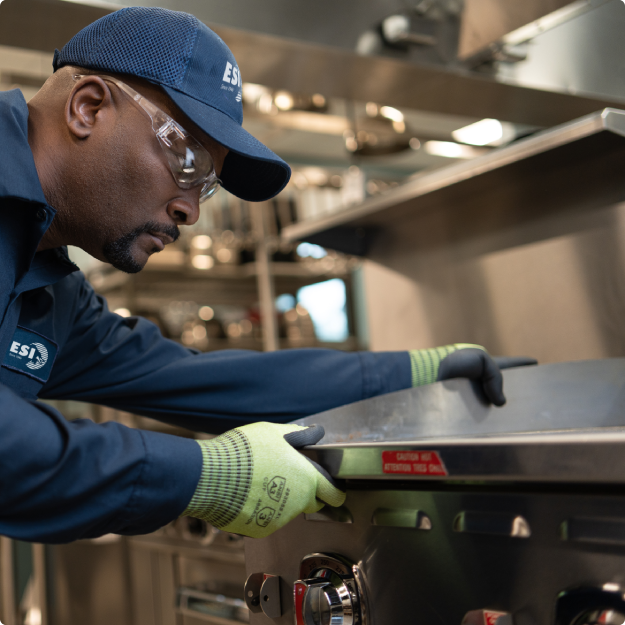 ESI technician in a branded navy work shirt and cap reaching across a stainless steel commercial range during a service call