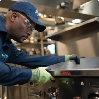 ESI technician in a branded navy work shirt and cap reaching across a stainless steel commercial range during a service call